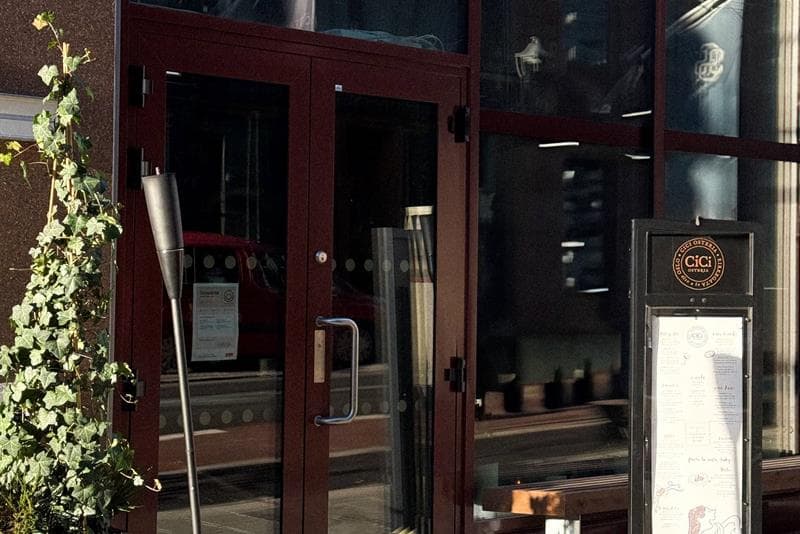 The entrance to a restaurant with large glass windows, a menu display by the door, and a lantern and plant placed on the pavement.