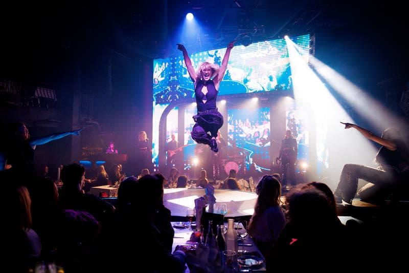 A performer leaps dramatically into the air before an illuminated audience as the stage glows in blue light. The backdrop features a live band and lig